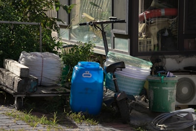 A cluttered outdoor area with various objects. There is a blue plastic drum, a bundle of white rope, a scooter leaned against some green baskets, and a green plastic trash can. Nearby, there is an air conditioning unit and dense greenery.