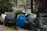 A cluttered outdoor area with various objects. There is a blue plastic drum, a bundle of white rope, a scooter leaned against some green baskets, and a green plastic trash can. Nearby, there is an air conditioning unit and dense greenery.