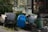 A cluttered outdoor area with various objects. There is a blue plastic drum, a bundle of white rope, a scooter leaned against some green baskets, and a green plastic trash can. Nearby, there is an air conditioning unit and dense greenery.