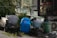 A cluttered outdoor area with various objects. There is a blue plastic drum, a bundle of white rope, a scooter leaned against some green baskets, and a green plastic trash can. Nearby, there is an air conditioning unit and dense greenery.