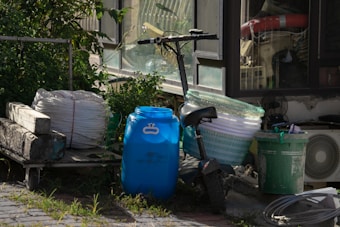A cluttered outdoor area with various objects. There is a blue plastic drum, a bundle of white rope, a scooter leaned against some green baskets, and a green plastic trash can. Nearby, there is an air conditioning unit and dense greenery.