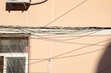 A beige wall features a window with an intricate metal grill. Numerous black cables criss-cross the scene, creating an elaborate web-like pattern. An air conditioning unit is mounted on the upper section of the wall.