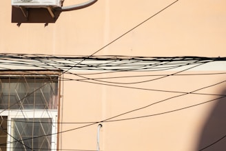 A beige wall features a window with an intricate metal grill. Numerous black cables criss-cross the scene, creating an elaborate web-like pattern. An air conditioning unit is mounted on the upper section of the wall.