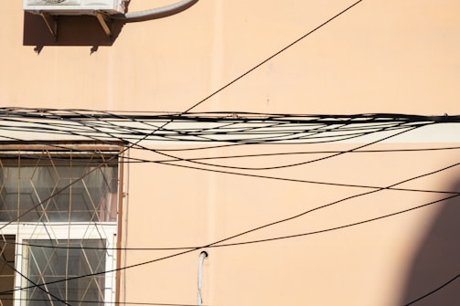 A beige wall features a window with an intricate metal grill. Numerous black cables criss-cross the scene, creating an elaborate web-like pattern. An air conditioning unit is mounted on the upper section of the wall.