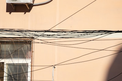 A beige wall features a window with an intricate metal grill. Numerous black cables criss-cross the scene, creating an elaborate web-like pattern. An air conditioning unit is mounted on the upper section of the wall.