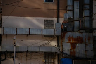 An urban scene featuring a building facade with various industrial elements. The wall is partially obscured by large ventilation ducts, some of which are rusted. Numerous wires and cables are crisscrossing in front of the structure. A security camera is mounted on a pole, indicating surveillance.