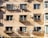 A beige apartment building facade features multiple windows with attached balconies. Each balcony houses small potted plants or clothes drying. Several air conditioning units are affixed to the wall, casting shadows in the evening light. The building's exterior shows some signs of aging.