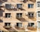 A beige apartment building facade features multiple windows with attached balconies. Each balcony houses small potted plants or clothes drying. Several air conditioning units are affixed to the wall, casting shadows in the evening light. The building's exterior shows some signs of aging.