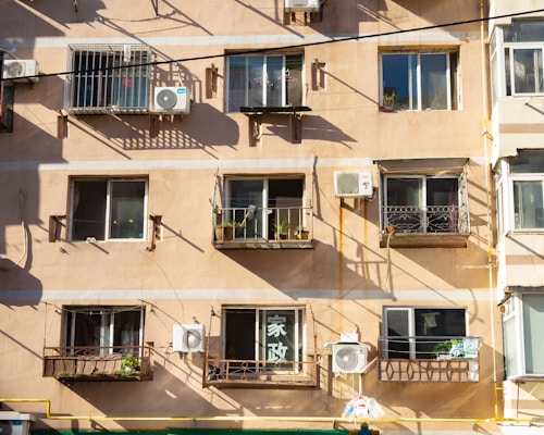 A beige apartment building facade features multiple windows with attached balconies. Each balcony houses small potted plants or clothes drying. Several air conditioning units are affixed to the wall, casting shadows in the evening light. The building's exterior shows some signs of aging.