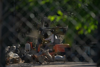 A person operates an orange forklift in an industrial setting, surrounded by various machinery parts and debris. The image is viewed through a chain-link fence, which adds a textured overlay to the scene. The surrounding environment is shaded by trees, casting a mix of light and shadow.