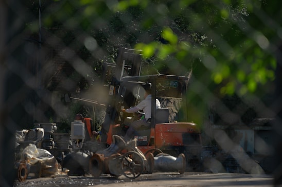 A person operates an orange forklift in an industrial setting, surrounded by various machinery parts and debris. The image is viewed through a chain-link fence, which adds a textured overlay to the scene. The surrounding environment is shaded by trees, casting a mix of light and shadow.