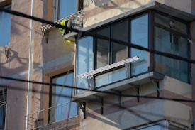 An external view of a building's upper floor with a glass-enclosed balcony. The balcony has metal railings and there are two ladders or railings stacked vertically. The building facade is beige with visible wear and tear. Electrical cables run across the foreground.