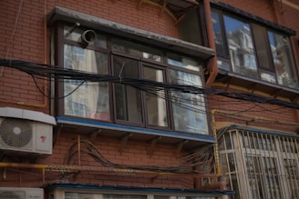 A brick building facade features several large windows with reflections of other structures. Numerous black cables and wires run horizontally across the exterior, accompanied by a wall-mounted air conditioning unit. The image conveys a sense of urban complexity and utility infrastructure.