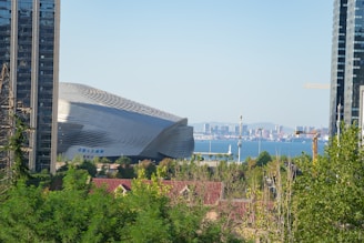 A modern architectural building with a sleek, futuristic design stands prominently among urban elements. In the background, a large body of water is visible with a clear skyline and distant city structures. The foreground includes green foliage, construction cranes, and surrounding high-rise buildings.