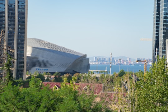 A modern architectural building with a sleek, futuristic design stands prominently among urban elements. In the background, a large body of water is visible with a clear skyline and distant city structures. The foreground includes green foliage, construction cranes, and surrounding high-rise buildings.