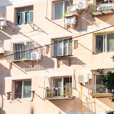 Several apartment windows and balconies are visible on a sunlit building facade. Air conditioning units are installed below some windows, and balconies have small plants and laundry items. Shadows of the railings and units are cast on the light peach-colored wall.