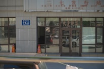 A building entrance with glass doors and windows, displaying various characters and signs in another language. Two orange traffic cones are placed near the entrance, and there is a blue address sign on the wall. The reflection of cars and street scenes is visible in the glass.