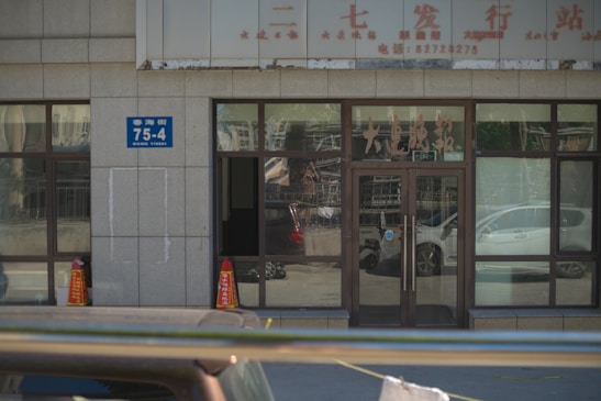A building entrance with glass doors and windows, displaying various characters and signs in another language. Two orange traffic cones are placed near the entrance, and there is a blue address sign on the wall. The reflection of cars and street scenes is visible in the glass.