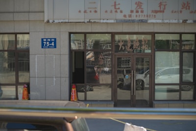 A building entrance with glass doors and windows, displaying various characters and signs in another language. Two orange traffic cones are placed near the entrance, and there is a blue address sign on the wall. The reflection of cars and street scenes is visible in the glass.