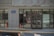 A building entrance with glass doors and windows, displaying various characters and signs in another language. Two orange traffic cones are placed near the entrance, and there is a blue address sign on the wall. The reflection of cars and street scenes is visible in the glass.