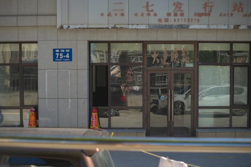 A building entrance with glass doors and windows, displaying various characters and signs in another language. Two orange traffic cones are placed near the entrance, and there is a blue address sign on the wall. The reflection of cars and street scenes is visible in the glass.