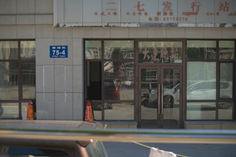 A building entrance with glass doors and windows, displaying various characters and signs in another language. Two orange traffic cones are placed near the entrance, and there is a blue address sign on the wall. The reflection of cars and street scenes is visible in the glass.