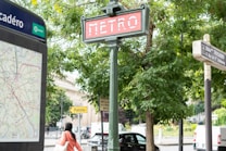 A vintage-style metro sign is prominently displayed in an outdoor urban environment, surrounded by lush green trees. A nearby map and directional signs are visible, and there is a person walking by with a bag over their shoulder. The scene captures an area bustling with transit and pedestrian activity.