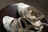 A pair of elegant, glittery high-heeled sandals with crisscross straps and ankle buckles resting on a dark wooden surface.
