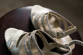 A pair of elegant, glittery high-heeled sandals with crisscross straps and ankle buckles resting on a dark wooden surface.