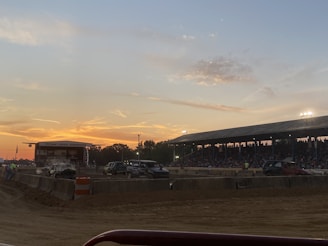 The scene captures a lively fairground with a large crowd gathered under a covered grandstand, watching an event on a dirt track. Several vehicles, including damaged or modified cars, are present on the track. The sky displays a colorful sunset, adding a warm glow to the environment.