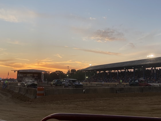 The scene captures a lively fairground with a large crowd gathered under a covered grandstand, watching an event on a dirt track. Several vehicles, including damaged or modified cars, are present on the track. The sky displays a colorful sunset, adding a warm glow to the environment.