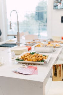 A bright kitchen setting with a white countertop featuring a platter of appetizers, possibly chicken wings, garnished with greens. Nearby, there are pink-colored pamphlets, and in the background, a modern sink faucet and other kitchen items are visible. Large windows allow plenty of natural light.