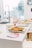 A bright kitchen setting with a white countertop featuring a platter of appetizers, possibly chicken wings, garnished with greens. Nearby, there are pink-colored pamphlets, and in the background, a modern sink faucet and other kitchen items are visible. Large windows allow plenty of natural light.