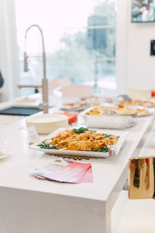 A bright kitchen setting with a white countertop featuring a platter of appetizers, possibly chicken wings, garnished with greens. Nearby, there are pink-colored pamphlets, and in the background, a modern sink faucet and other kitchen items are visible. Large windows allow plenty of natural light.