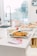 A bright kitchen setting with a white countertop featuring a platter of appetizers, possibly chicken wings, garnished with greens. Nearby, there are pink-colored pamphlets, and in the background, a modern sink faucet and other kitchen items are visible. Large windows allow plenty of natural light.