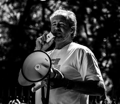 A man holding a megaphone and a microphone, seemingly speaking in an outdoor setting. The image is in black and white, with dappled sunlight creating contrasting shadows on the man's face and clothes.