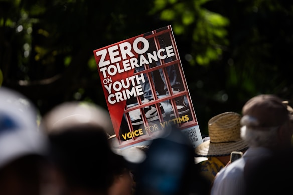A protest sign held in a crowd with the text 'ZERO TOLERANCE ON YOUTH CRIME' displayed. The sign features an image of a young person behind bars, highlighting a message for victims. The background shows people gathered, some wearing hats, surrounded by greenery, suggesting the event is outdoors.