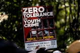 A protest sign featuring bold white letters on a red background with the message 'Zero Tolerance on Youth Crime.' The sign shows the image of legs in sneakers behind bars, symbolizing imprisonment. The phrase 'Voice for Victims' is written at the bottom in yellow.
