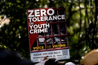 A protest sign featuring bold white letters on a red background with the message 'Zero Tolerance on Youth Crime.' The sign shows the image of legs in sneakers behind bars, symbolizing imprisonment. The phrase 'Voice for Victims' is written at the bottom in yellow.