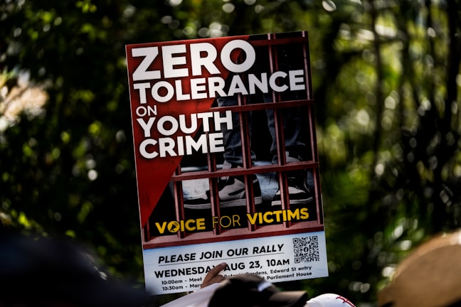 A protest sign featuring bold white letters on a red background with the message 'Zero Tolerance on Youth Crime.' The sign shows the image of legs in sneakers behind bars, symbolizing imprisonment. The phrase 'Voice for Victims' is written at the bottom in yellow.