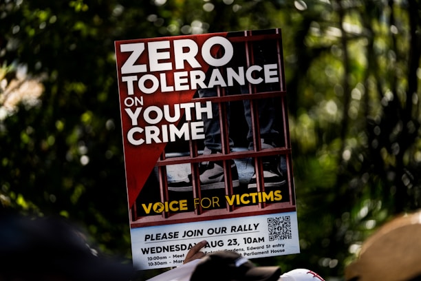 A protest sign featuring bold white letters on a red background with the message 'Zero Tolerance on Youth Crime.' The sign shows the image of legs in sneakers behind bars, symbolizing imprisonment. The phrase 'Voice for Victims' is written at the bottom in yellow.