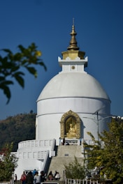 A large white stupa with a gold-topped spire is prominently positioned against a clear blue sky. A golden statue of Buddha is housed in an ornate alcove at the front. People can be seen walking towards the structure, which is surrounded by greenery and hills in the background.