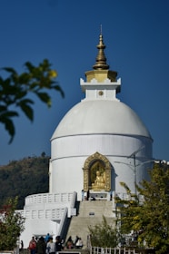 A large white stupa with a gold-topped spire is prominently positioned against a clear blue sky. A golden statue of Buddha is housed in an ornate alcove at the front. People can be seen walking towards the structure, which is surrounded by greenery and hills in the background.