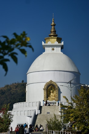 A large white stupa with a gold-topped spire is prominently positioned against a clear blue sky. A golden statue of Buddha is housed in an ornate alcove at the front. People can be seen walking towards the structure, which is surrounded by greenery and hills in the background.