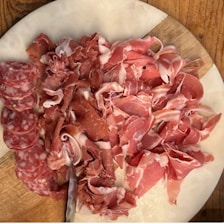 A wooden board with an assortment of cured meats including sliced salami, prosciutto, and other types of thinly sliced deli meats. The meats are arranged neatly on the board, showcasing a variety of red and pink hues with marbled fat.