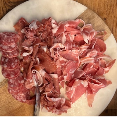 A wooden board with an assortment of cured meats including sliced salami, prosciutto, and other types of thinly sliced deli meats. The meats are arranged neatly on the board, showcasing a variety of red and pink hues with marbled fat.