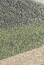 A close-up view of broken glass with a multitude of small, fragmented pieces creating an intricate and chaotic pattern. The glass reflects light, displaying a variety of textures and transparency levels, set against a blurred green and beige background.
