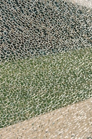 A close-up view of broken glass with a multitude of small, fragmented pieces creating an intricate and chaotic pattern. The glass reflects light, displaying a variety of textures and transparency levels, set against a blurred green and beige background.