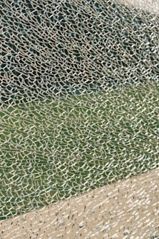 A close-up view of broken glass with a multitude of small, fragmented pieces creating an intricate and chaotic pattern. The glass reflects light, displaying a variety of textures and transparency levels, set against a blurred green and beige background.