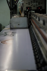 A large industrial machine standing in a factory environment is captured up-close. The machine appears to be a printer or fabric printing device. The conveyor belt on the machine has a visible pattern, and there are various mechanical parts visible, showing its operational complexities.